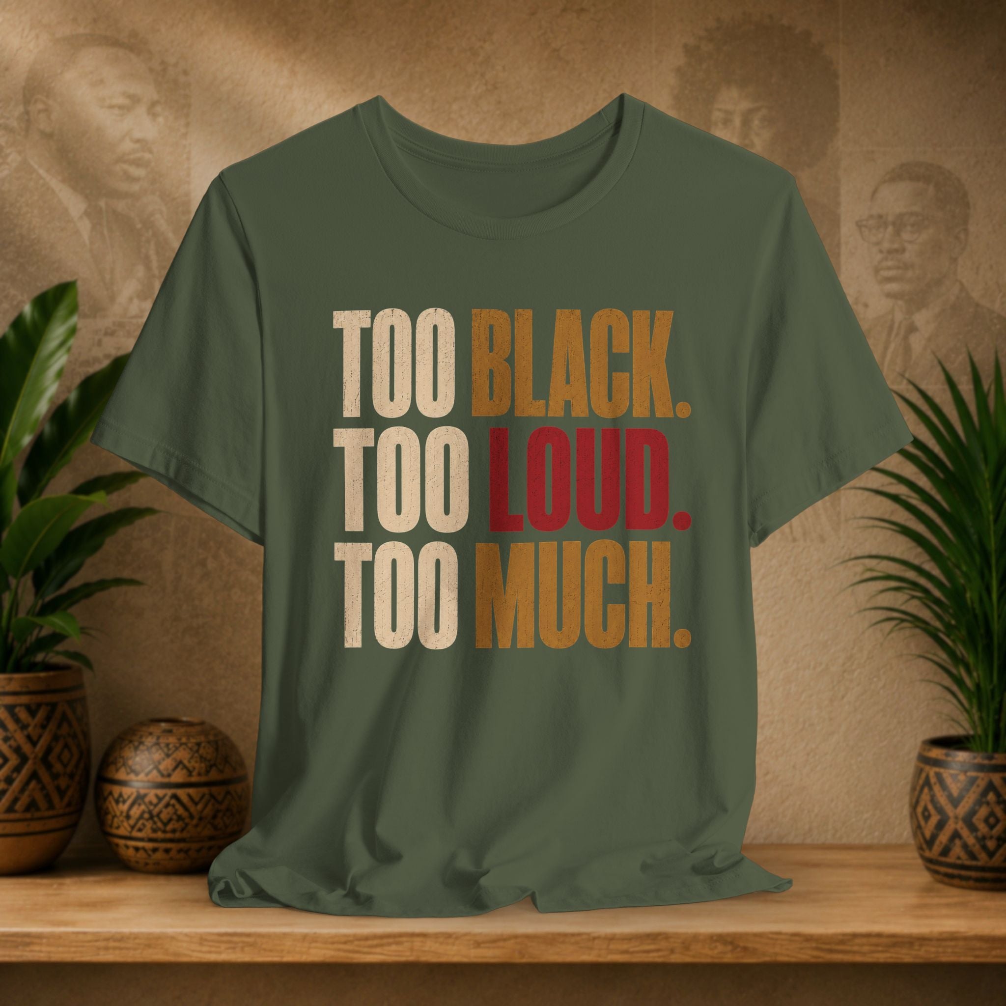 An olive green graphic t-shirt displayed on a wooden shelf. The shirt features the text 'TOO BLACK.
