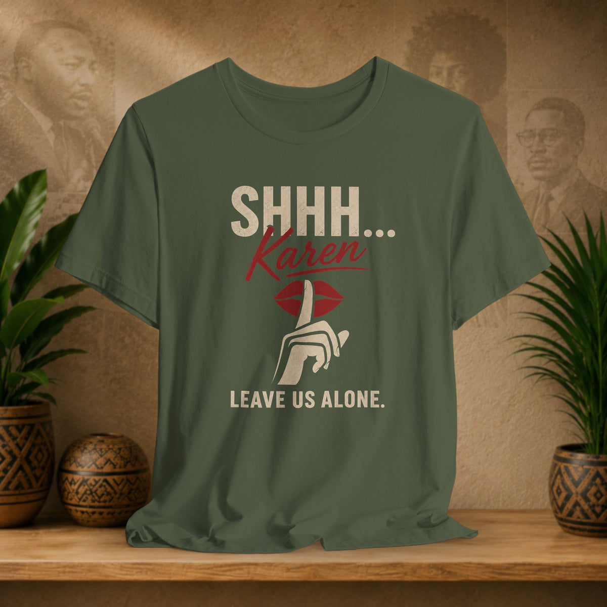 An olive green graphic t-shirt displayed on a wooden shelf. The shirt features a printed design with the text 'SHHH.