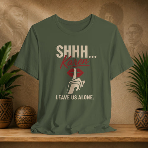 An olive green graphic t-shirt displayed on a wooden shelf. The shirt features a printed design with the text 'SHHH.