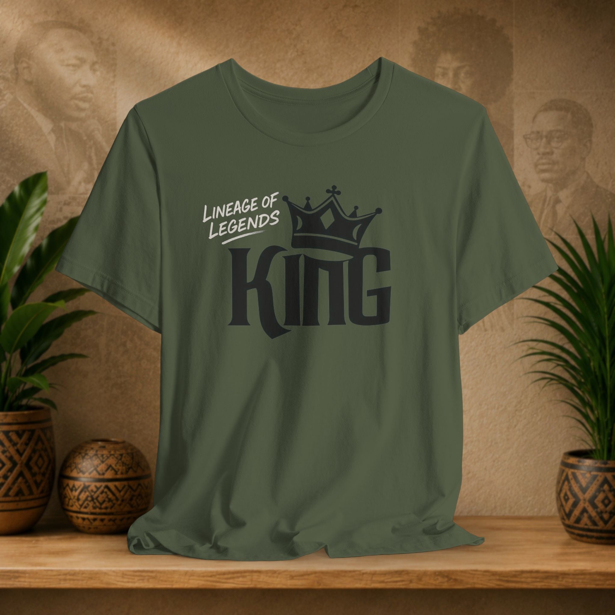Military green Lineage of Legends King graphic tee with black crown design, displayed on a shelf with plants and Afrocentric portraits