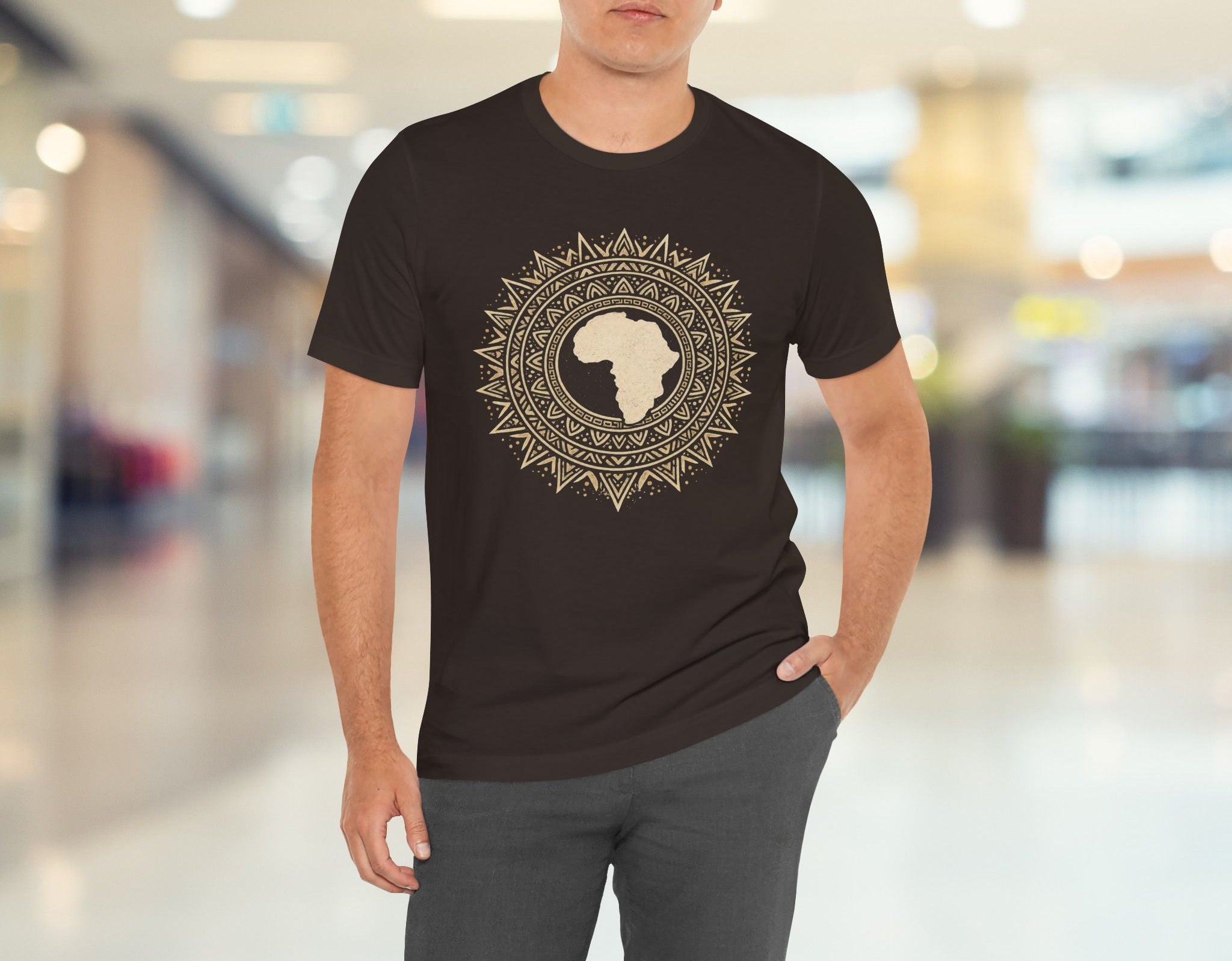 Man wearing a brown t-shirt with a circular design featuring a stylised map of Africa, standing in a blurred indoor setting.