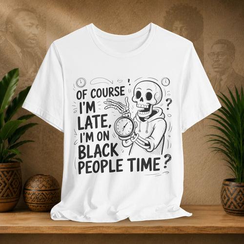 A white short-sleeve graphic T-shirt displayed on a wooden surface, featuring a black line-art design of a skeleton holding a clock with the text 'OF COURSE I'M LATE, I'M ON BLACK PEOPLE TIME?'. The shirt is styled in a mockup setting with potted plants and a textured brown background.