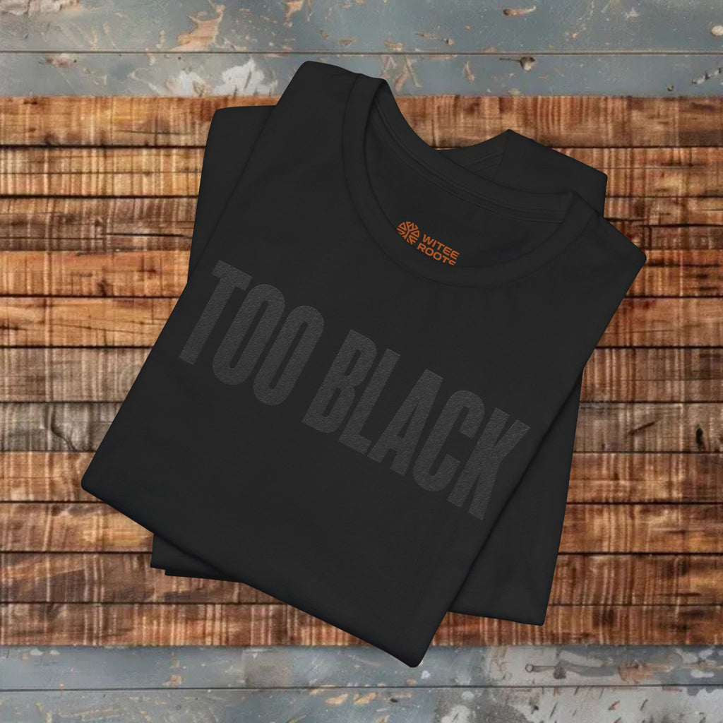 A folded black t-shirt with 'TOO BLACK' printed in a dark gray tone across the front, featuring a small orange 'WITNESWEEKS' logo inside the collar. The shirt is laid flat on a rustic wooden plank surface.
