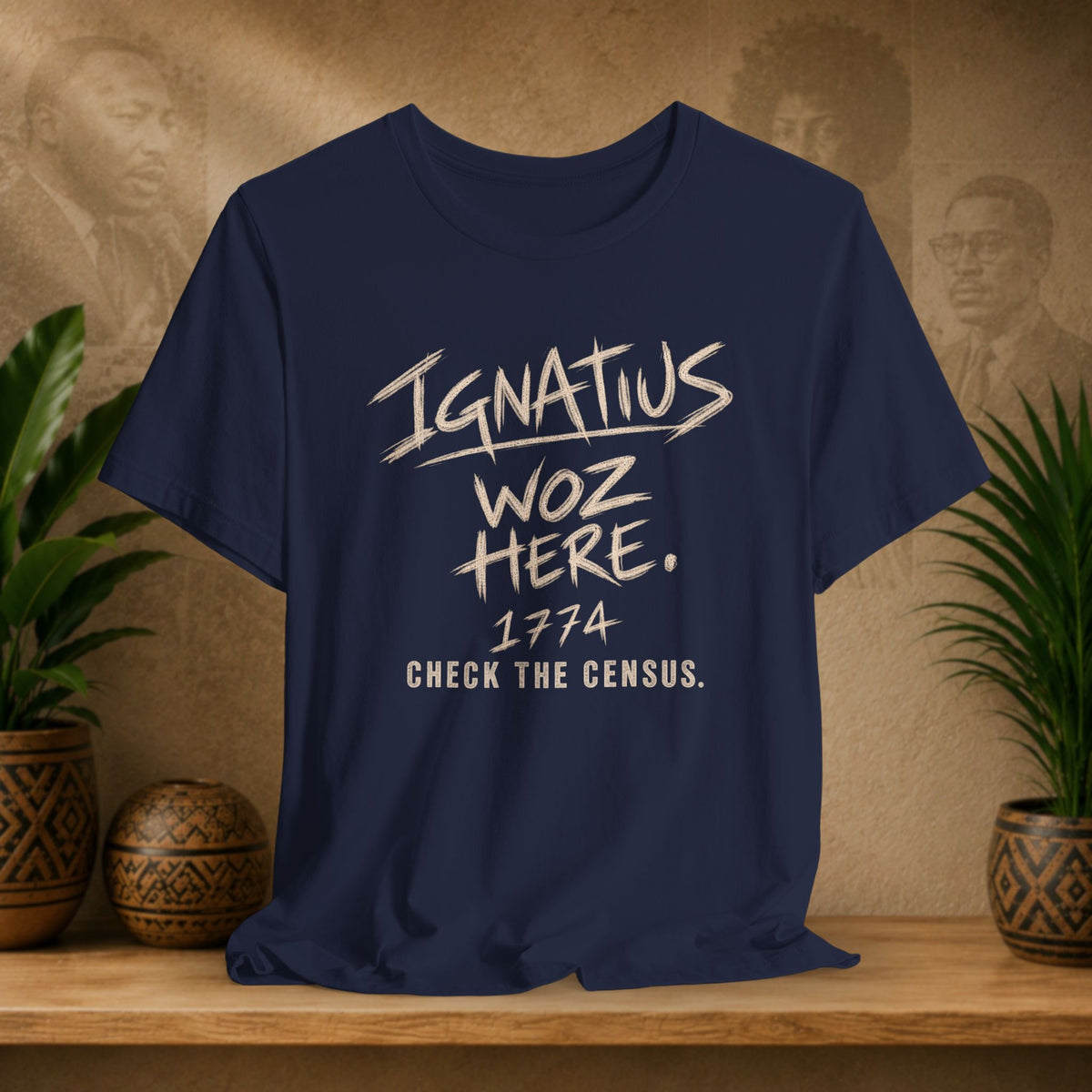 A navy blue graphic t-shirt displayed on a wooden shelf, flanked by potted plants and decorative vases against a textured beige background with sepia portraits.