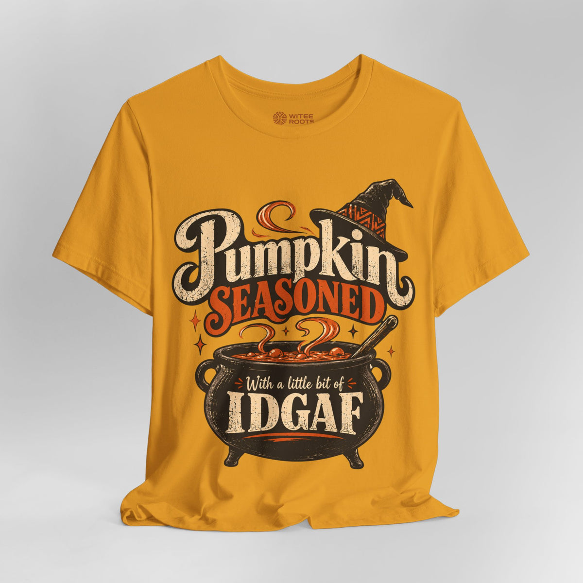 A mustard yellow t-shirt with a Halloween graphic that reads 'Pumpkin Seasoned With a little bit of IDGAF' above a cauldron and witch hat, shown on a light gray background.