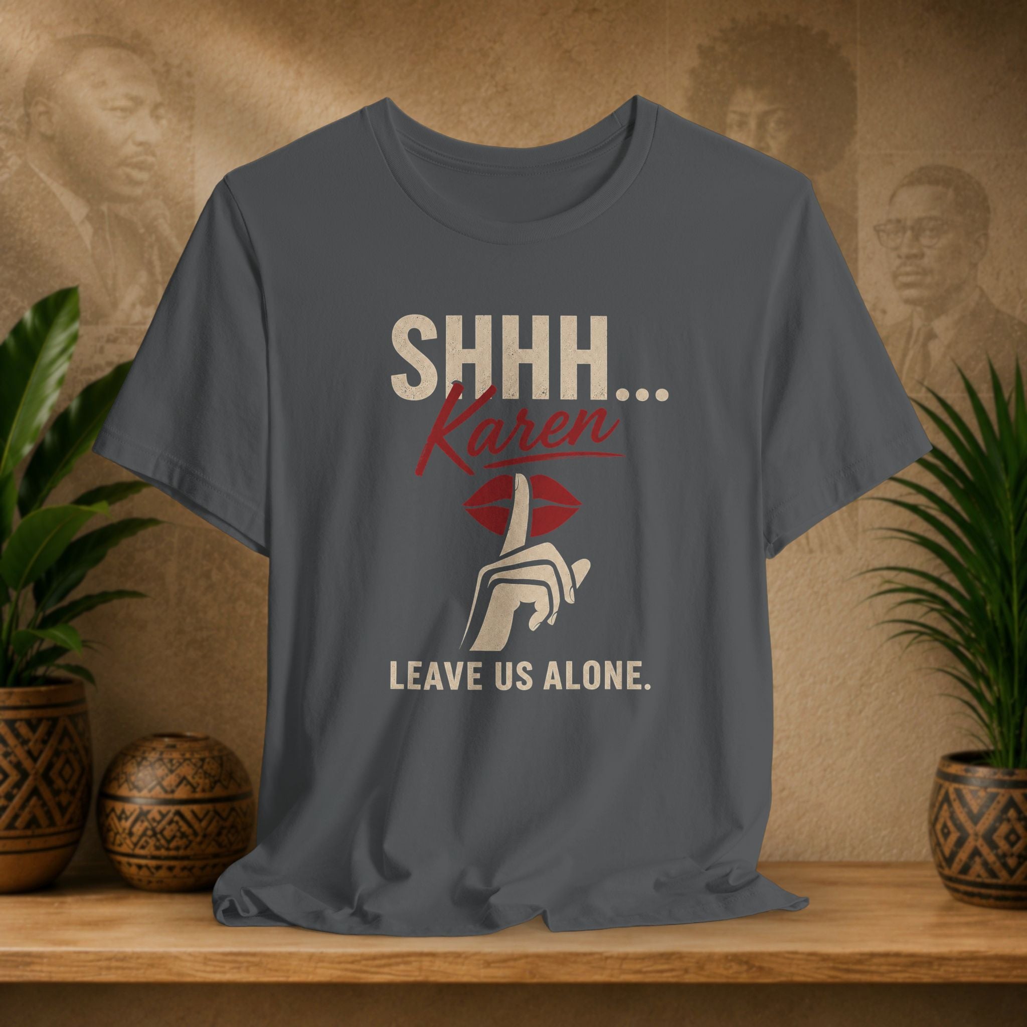 A dark gray graphic t-shirt displayed on a wooden shelf. The shirt features a graphic print with the text 'SHHH.