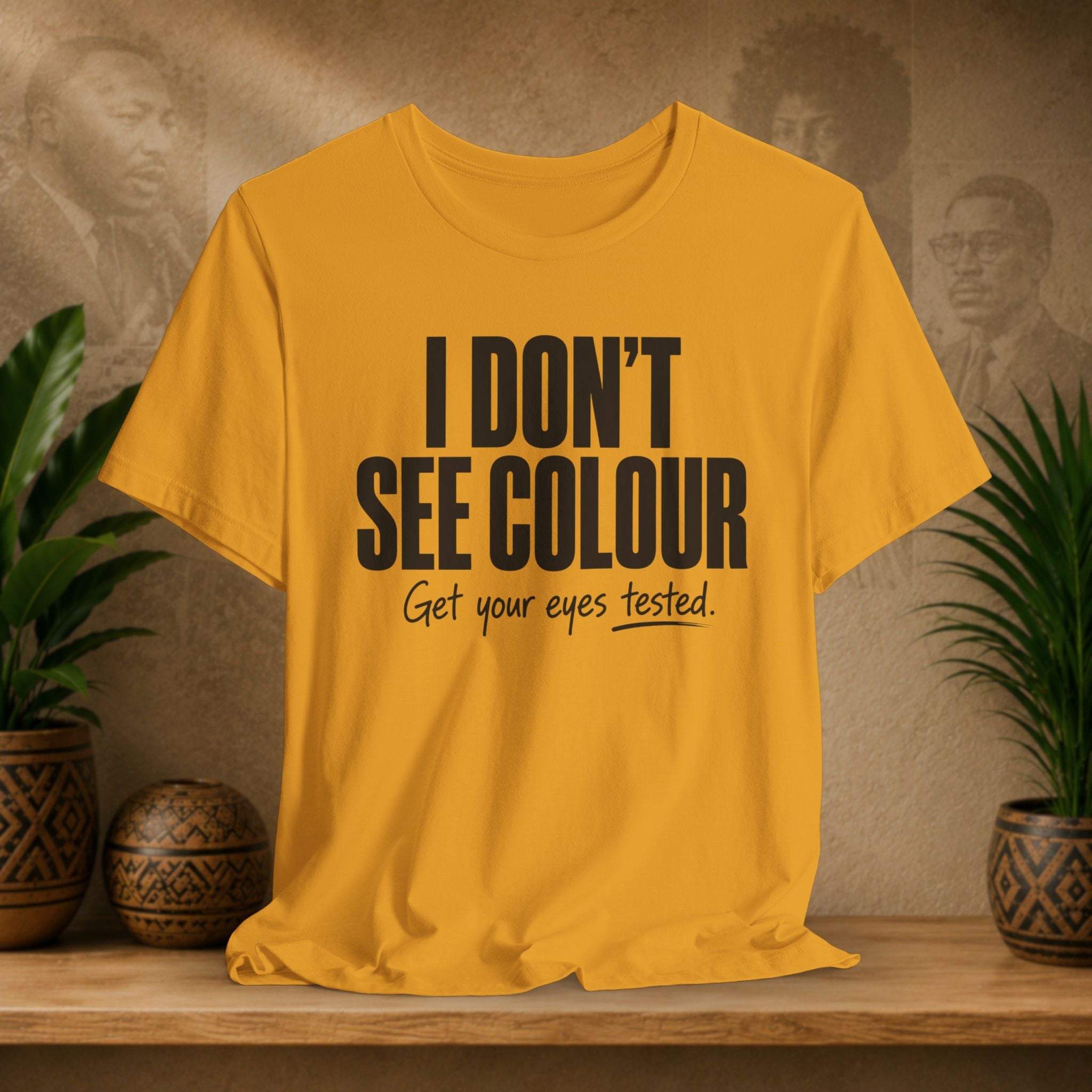Yellow t-shirt with 'I Don't See Colour' text on a wooden surface with plants and a textured wall background.
