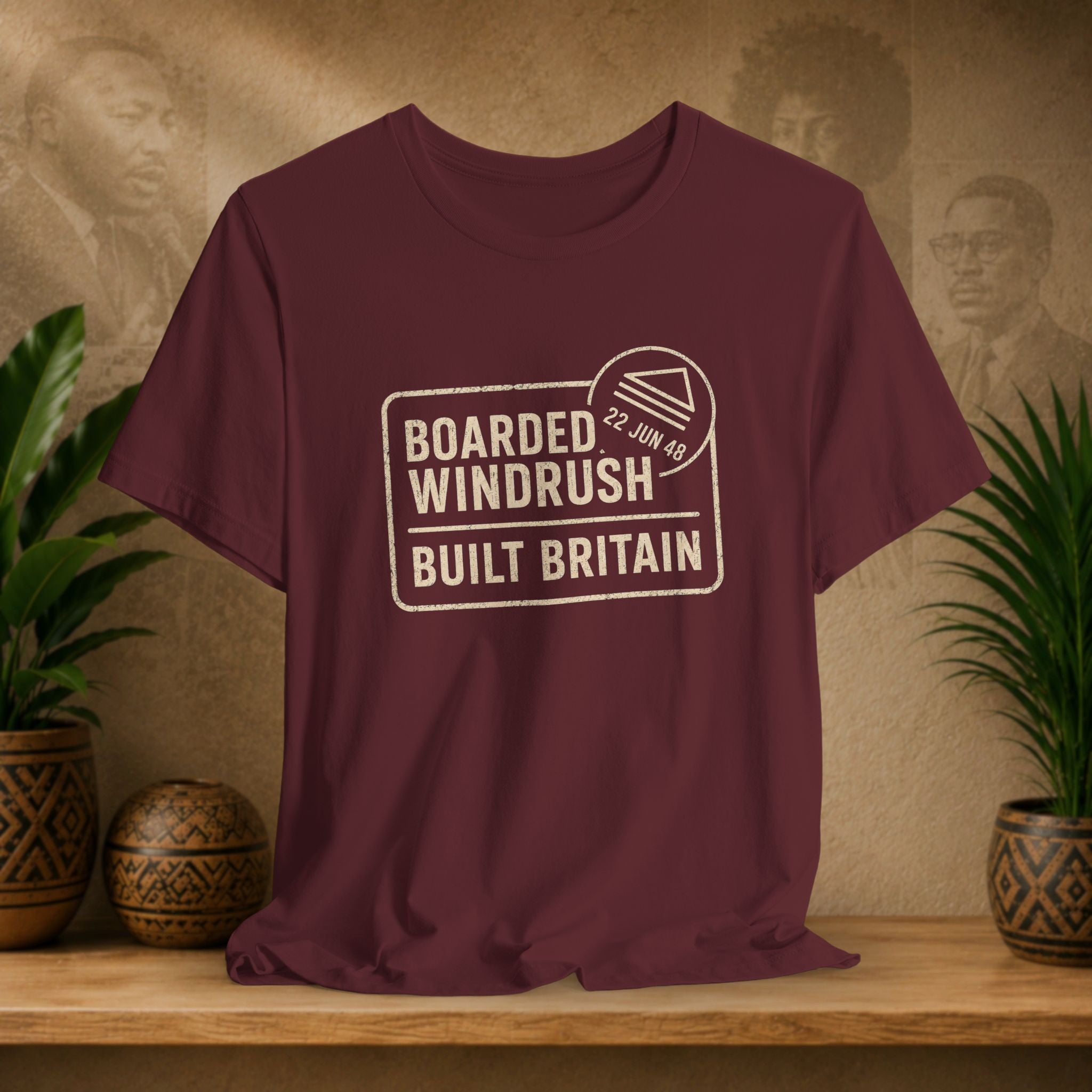 Maroon t-shirt with 'Boarded Windrush Built Britain' text on a wooden surface with plants and a textured wall background.