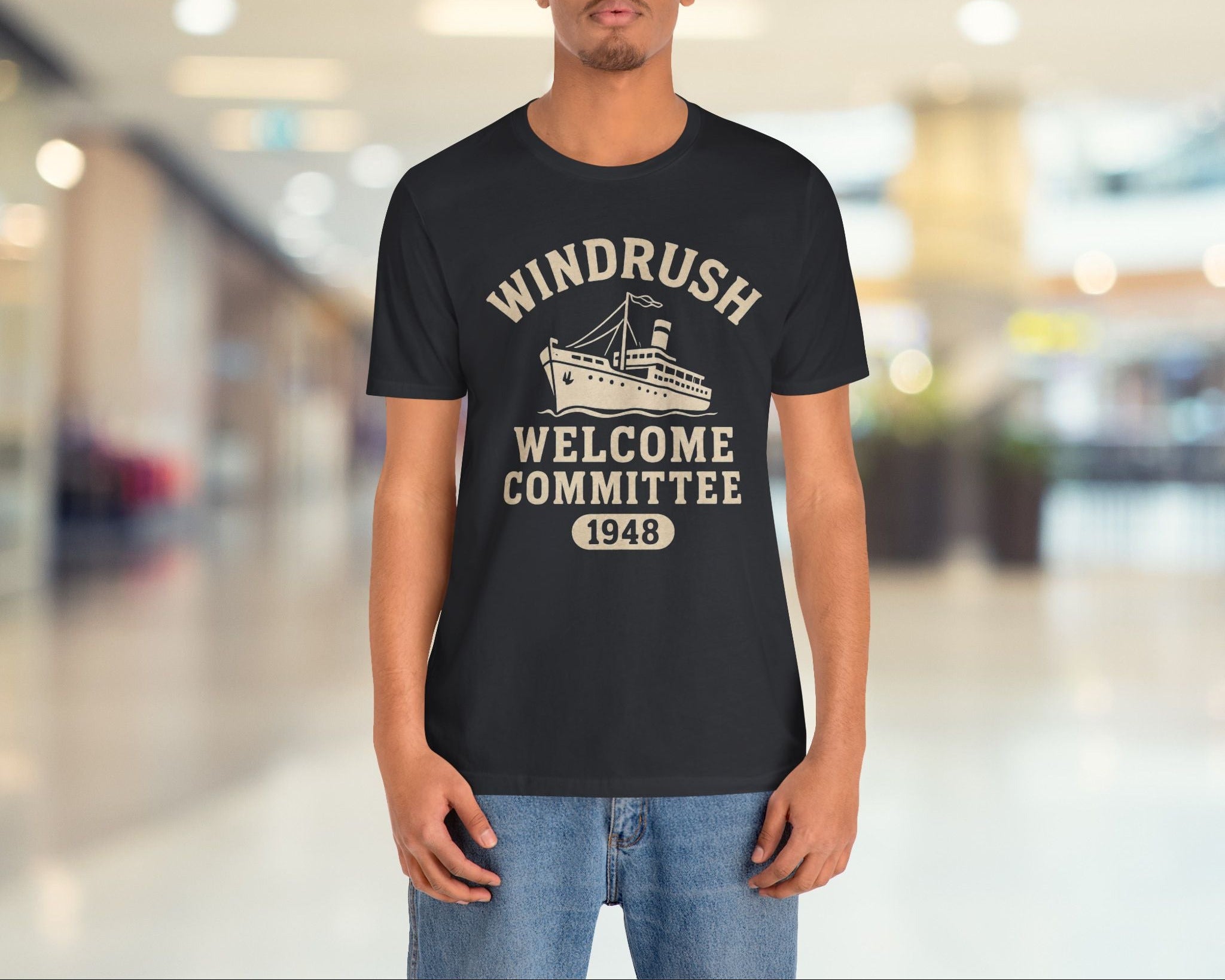 Person wearing a black t-shirt with 'Windrush Welcome Committee 1948' text and graphic in a blurred indoor setting.