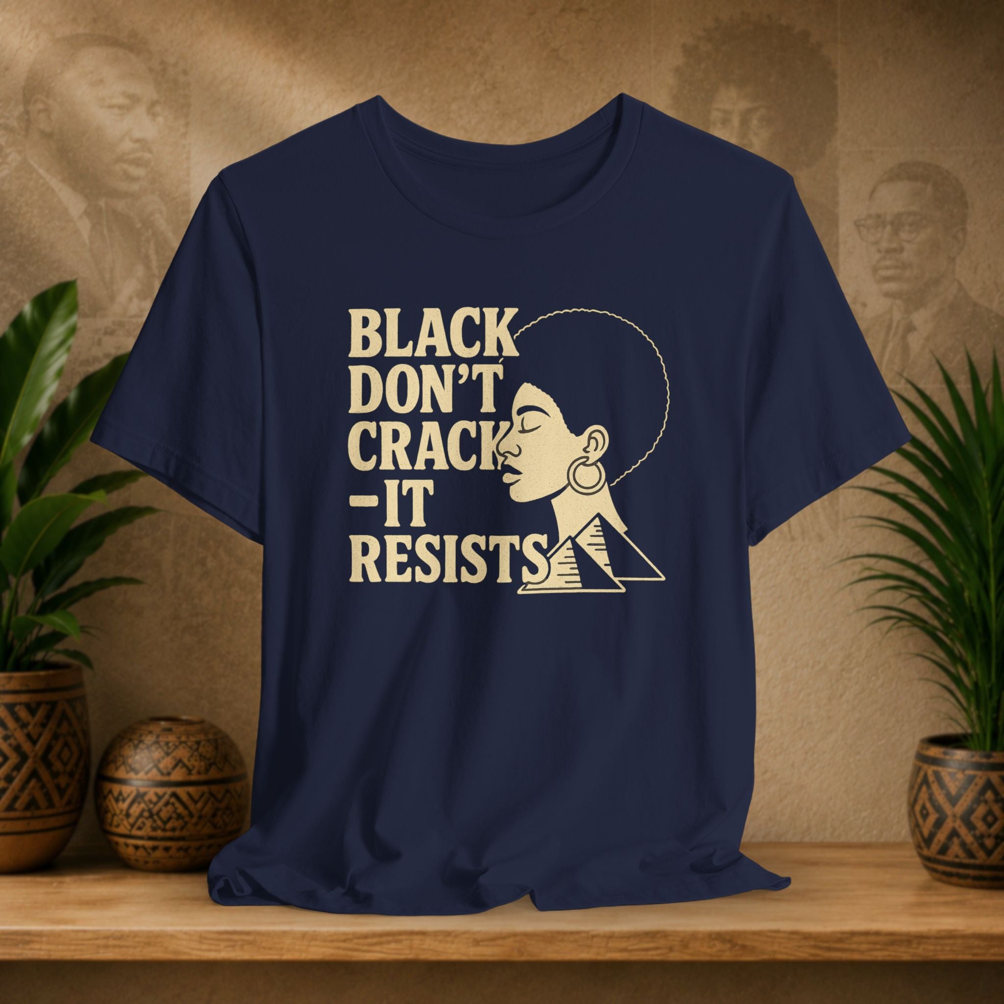 Navy blue t-shirt with 'Black Don't Crack, It Resists' text and graphic on a wooden surface with plants and decorative items.