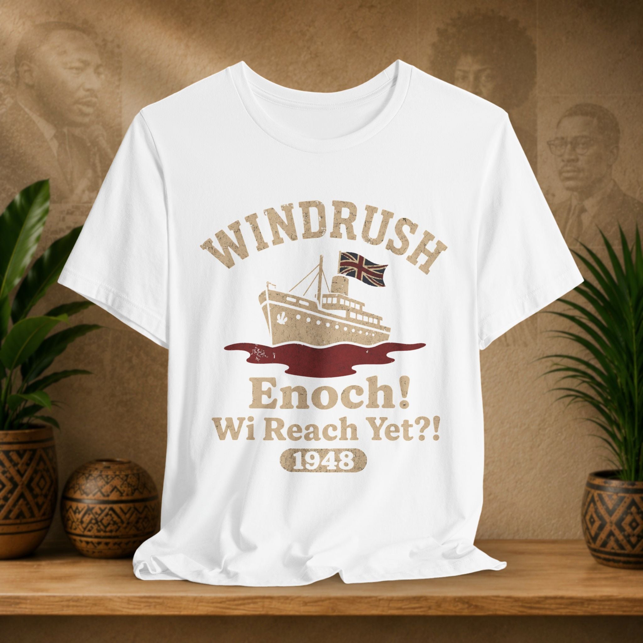 White t-shirt with 'Windrush Enoch Wi Reach Yet?' design on a wooden surface with plants and a textured wall background.