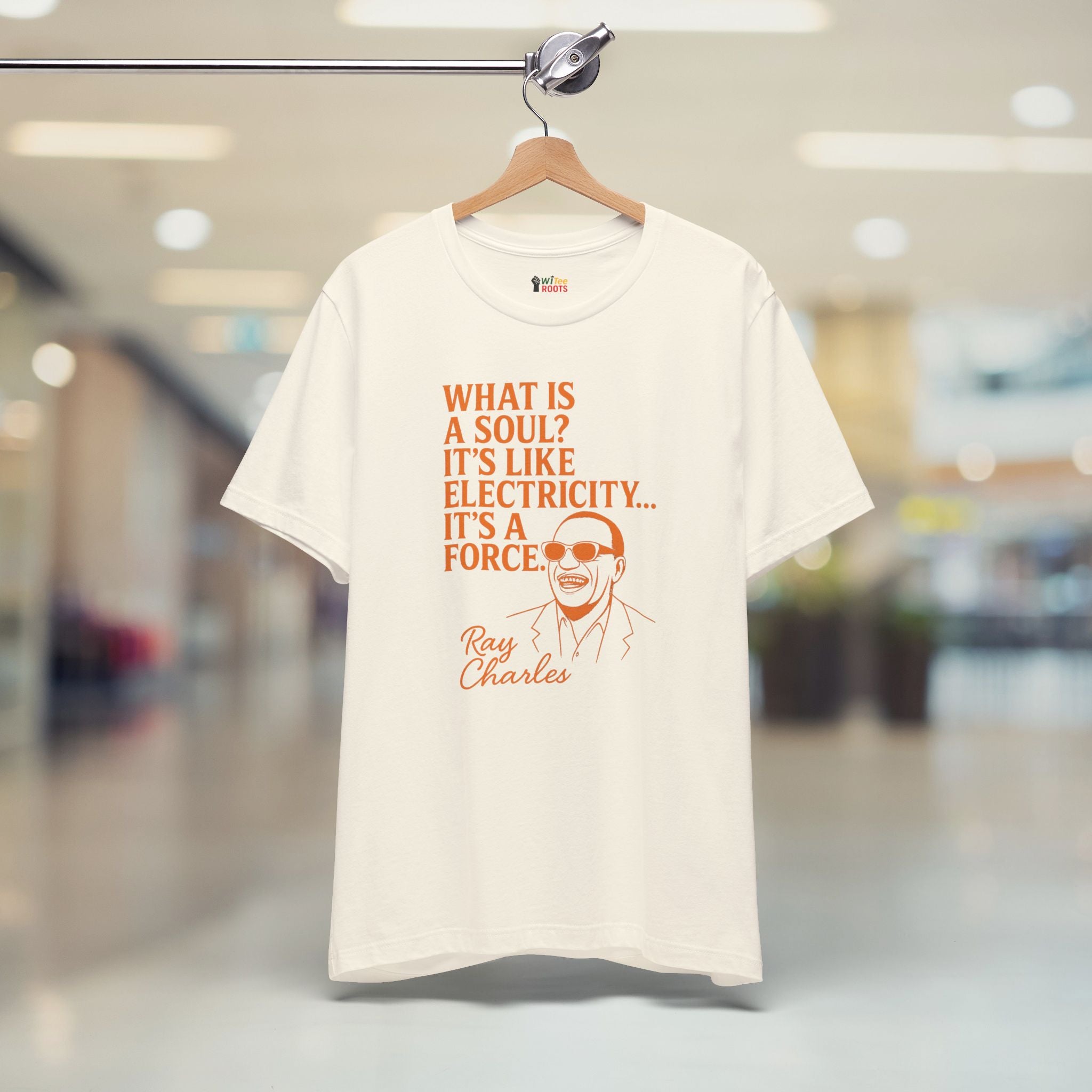 Beige t-shirt with orange text and graphic of Ray Charles on a hanger in a store.