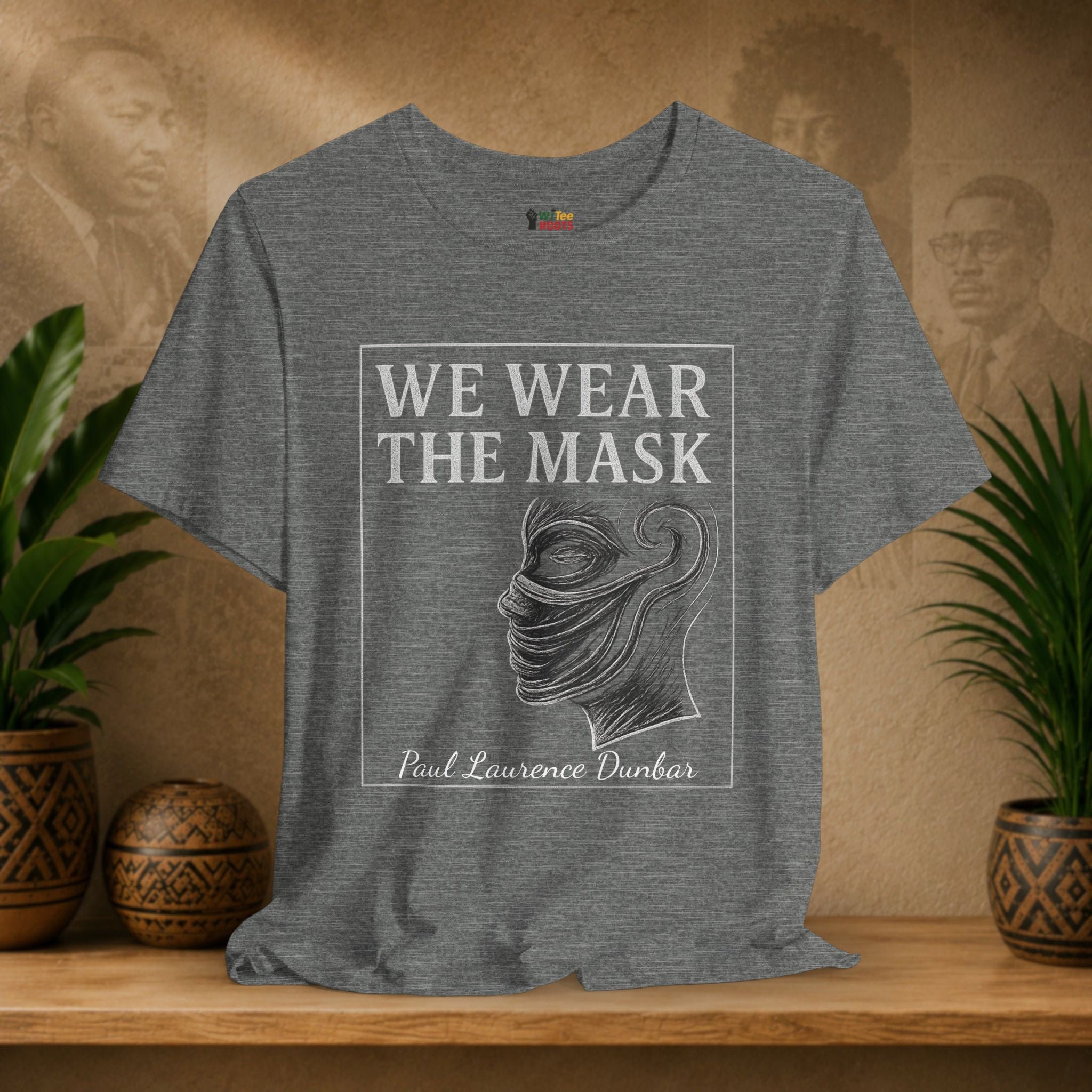 Gray t-shirt with 'We Wear the Mask' quote by Paul Laurence Dunbar on a wooden shelf with plants and decorative items.