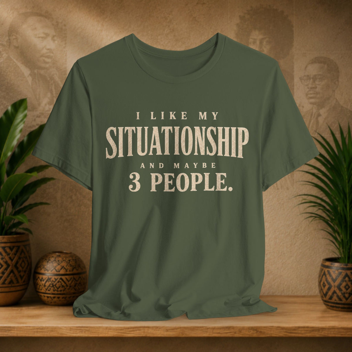 An olive green short-sleeve t-shirt with a cream-colored text graphic reading 'I LIKE MY SITUATIONSHIP AND MAYBE 3 PEOPLE.' displayed on a wooden shelf with potted plants and a textured background.