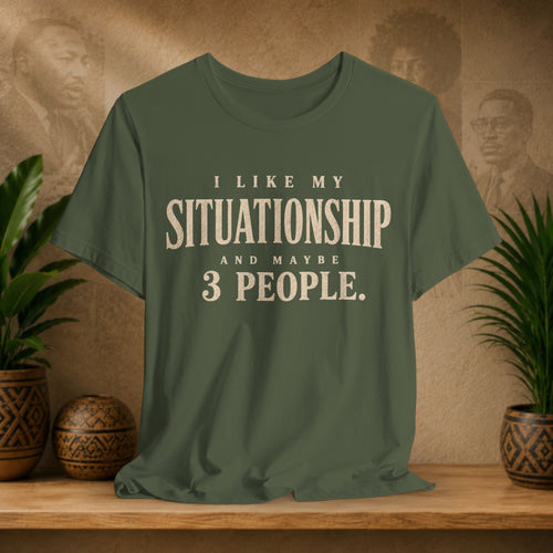 An olive green short-sleeve t-shirt with a cream-colored text graphic reading 'I LIKE MY SITUATIONSHIP AND MAYBE 3 PEOPLE.' displayed on a wooden shelf with potted plants and a textured background.
