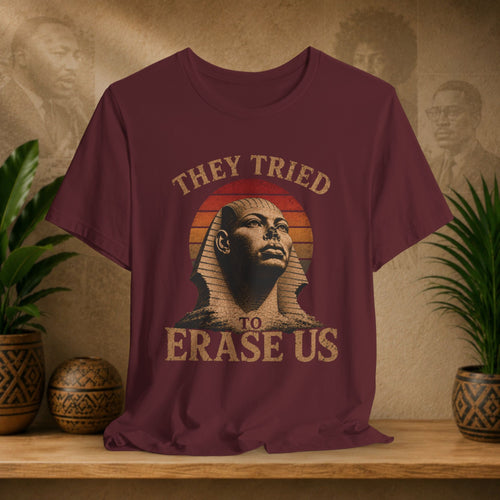 Maroon t-shirt with a graphic of a sphinx and text 'They tried to erase us' on a wooden surface with plants and a textured wall in the background.