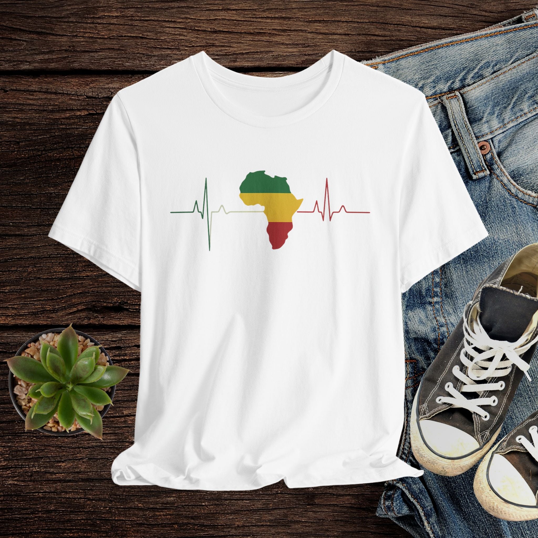 White t-shirt with a colorful Africa map design on a wooden surface with jeans and sneakers.