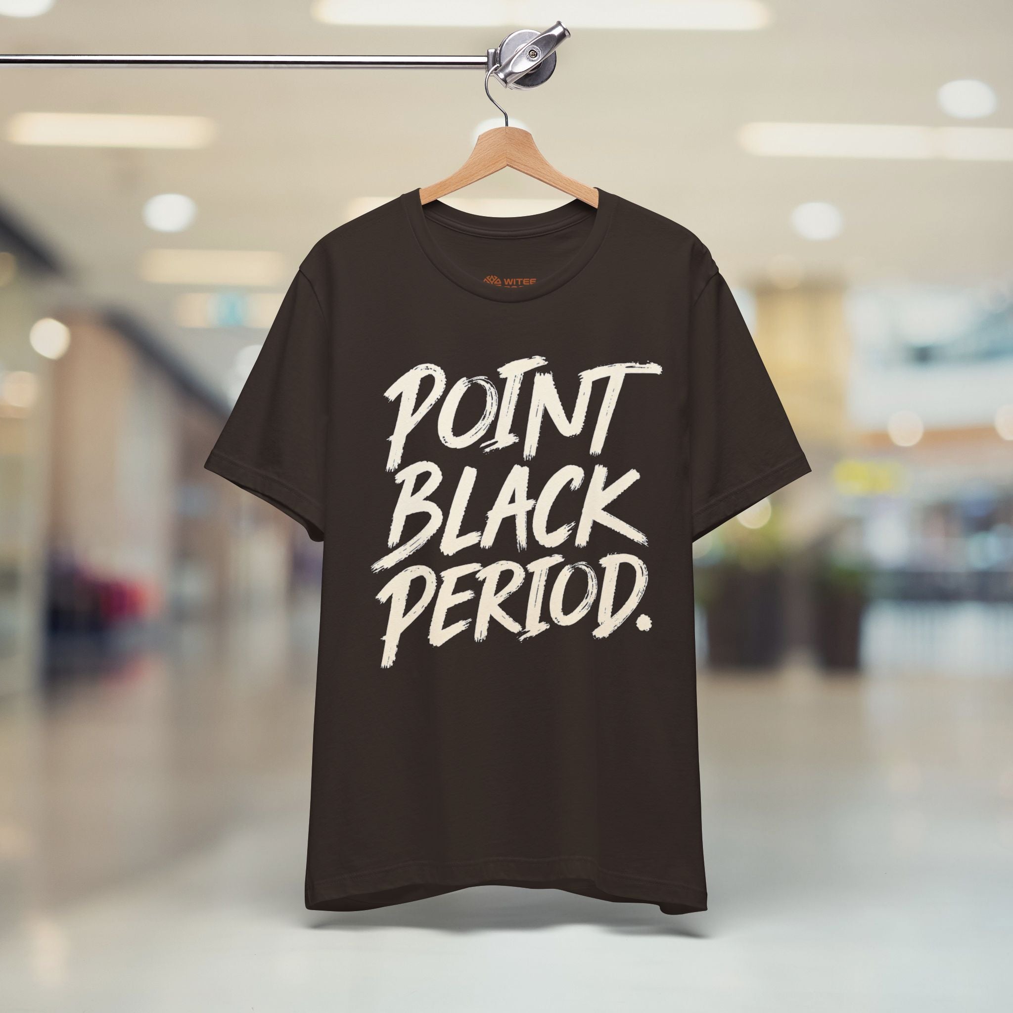A dark brown short-sleeve t-shirt hanging on a wooden hanger from a clothing rack, featuring the text 'POINT BLACK PERIOD.' in a white brush-stroke font on the front.