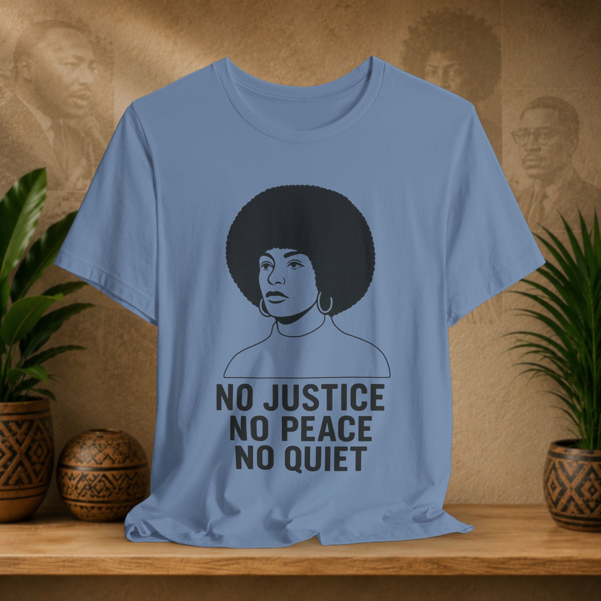 Blue t-shirt with a graphic of a person with an afro and text 'No Justice No Peace No Quiet' on a wooden surface with plants and a textured wall in the background.