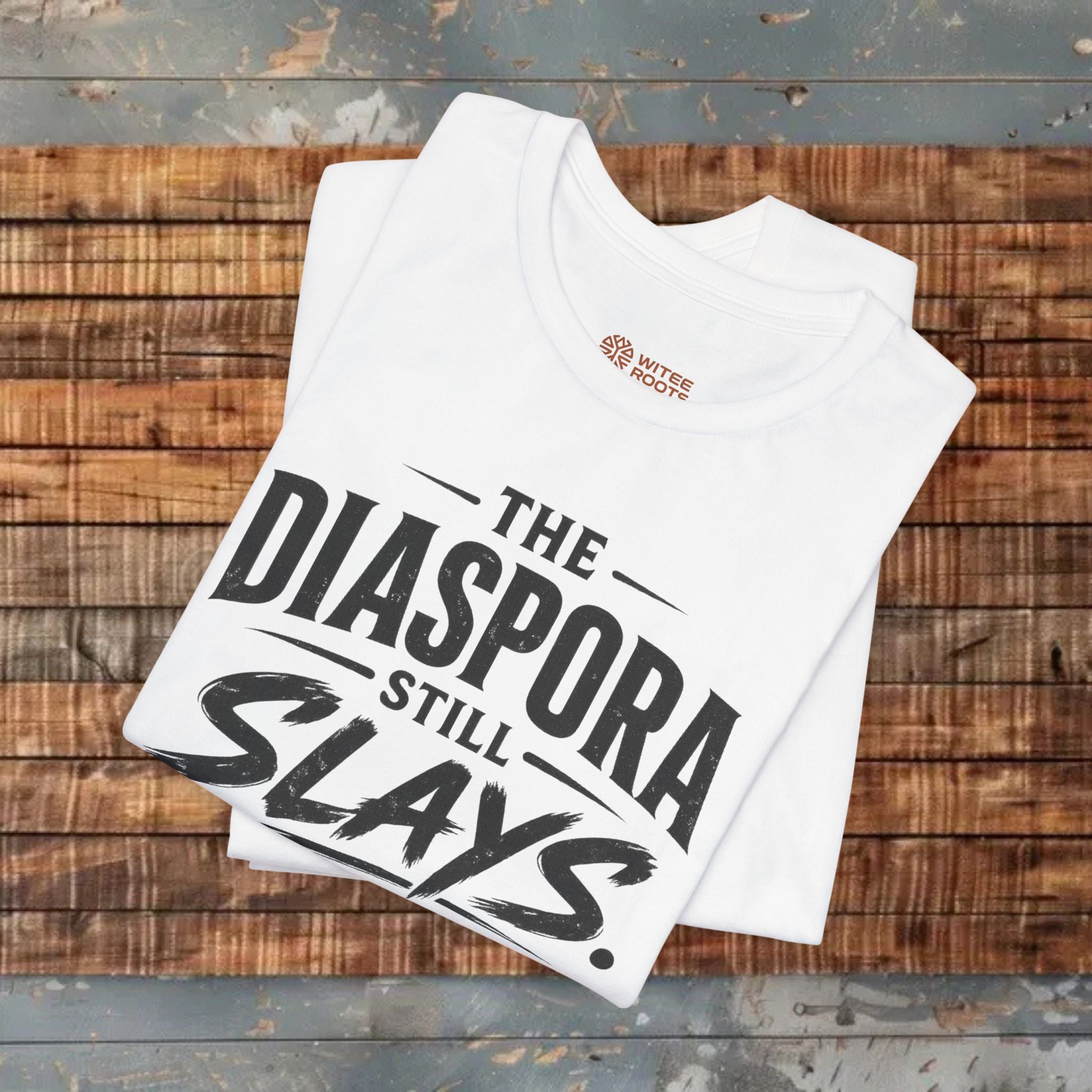 White folded t-shirt with 'The Diaspora Still Slays Every Generation' text on a wooden background.