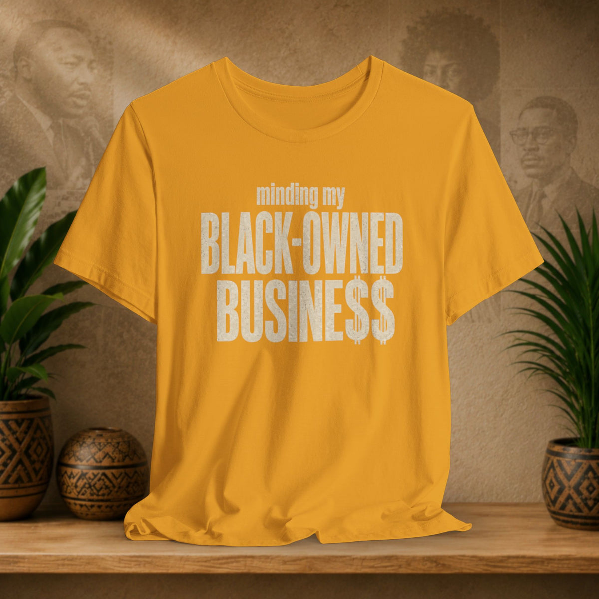 A mustard yellow t-shirt with a distressed white graphic reading 'minding my BLACK-OWNED BUSINESS' displayed on a wooden shelf with potted plants and a textured background.