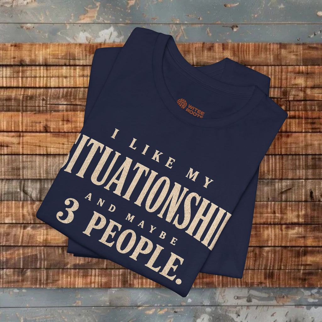 A navy blue folded t-shirt with a humorous beige graphic print reading 'I LIKE MY SITUATIONS AND MAYBE 3 PEOPLE.' displayed on a rustic wooden plank surface.