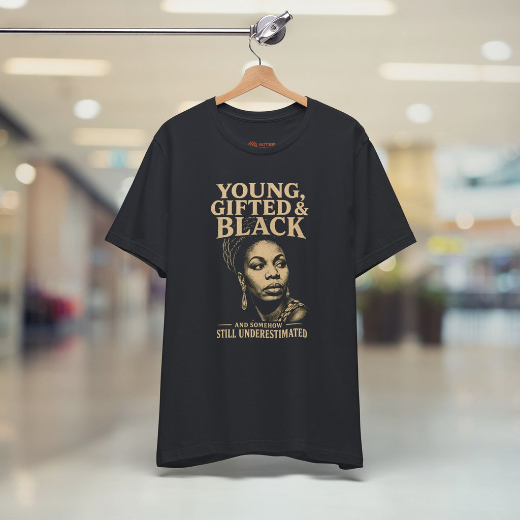 graphic t-shirt with a gold and white print that reads 'YOUNG, GIFTED & BLACK' and 'JEN NASHOR IS STILL UNDERESTIMATED', standing in a blurred indoor hallway.