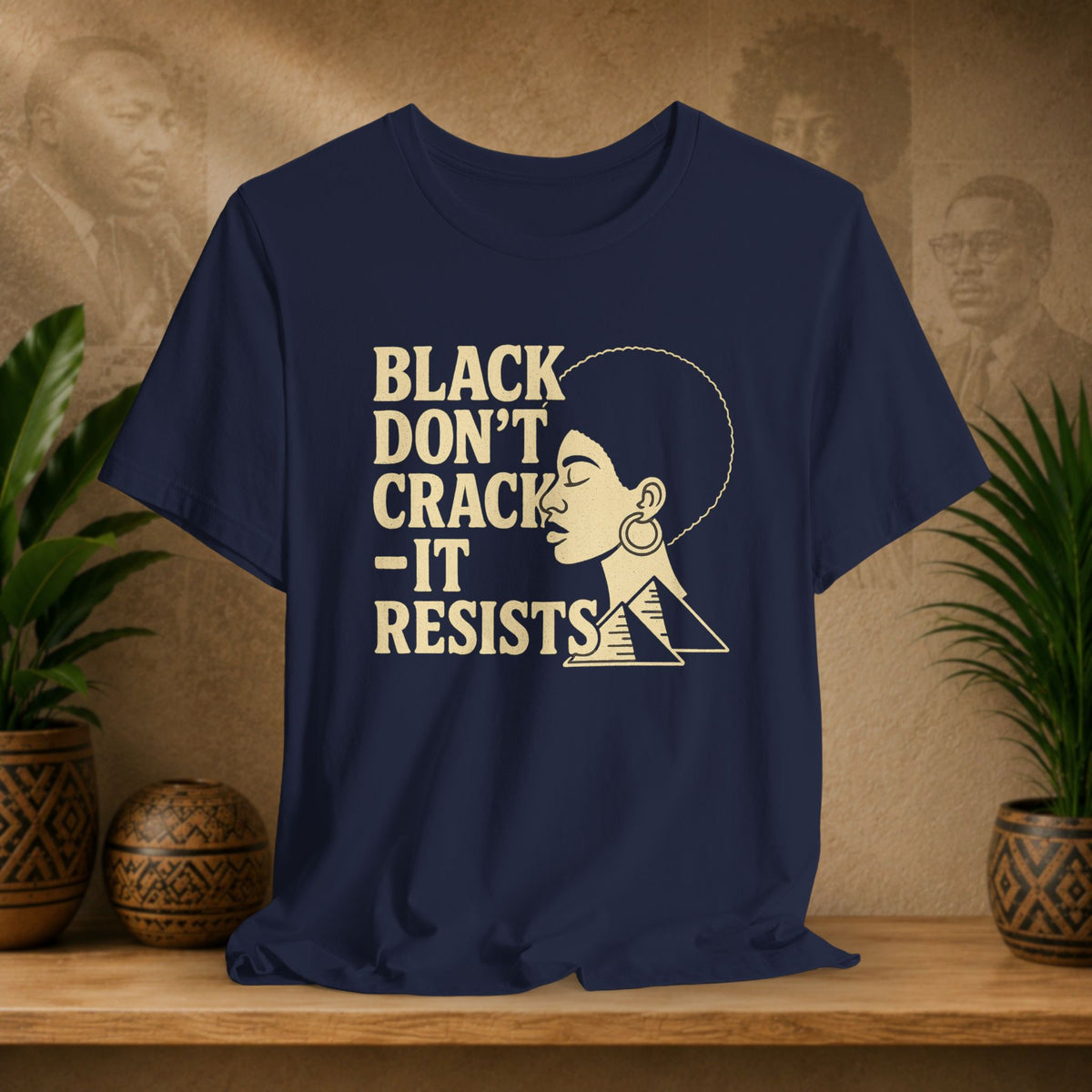 A navy blue graphic t-shirt displayed on a wooden shelf, flanked by potted plants. The shirt features a cream-colored graphic of a woman's profile next to the text 'BLACK DON'T CRACK - IT RESISTS'.