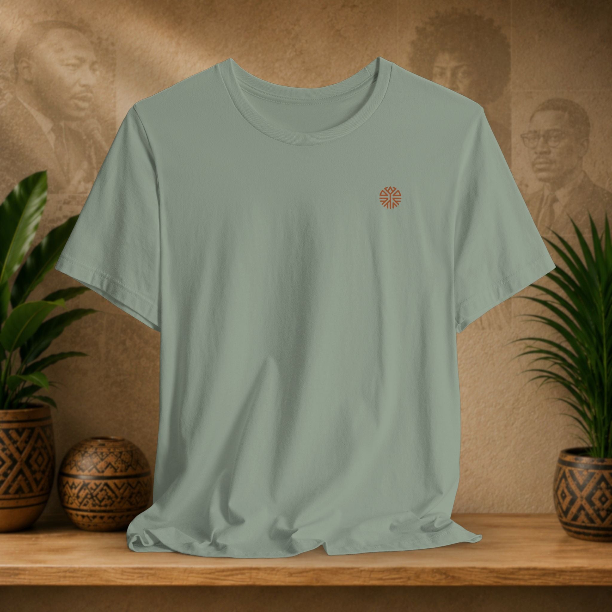 Sage green t-shirt with a small logo on a wooden surface with plants and a textured wall background