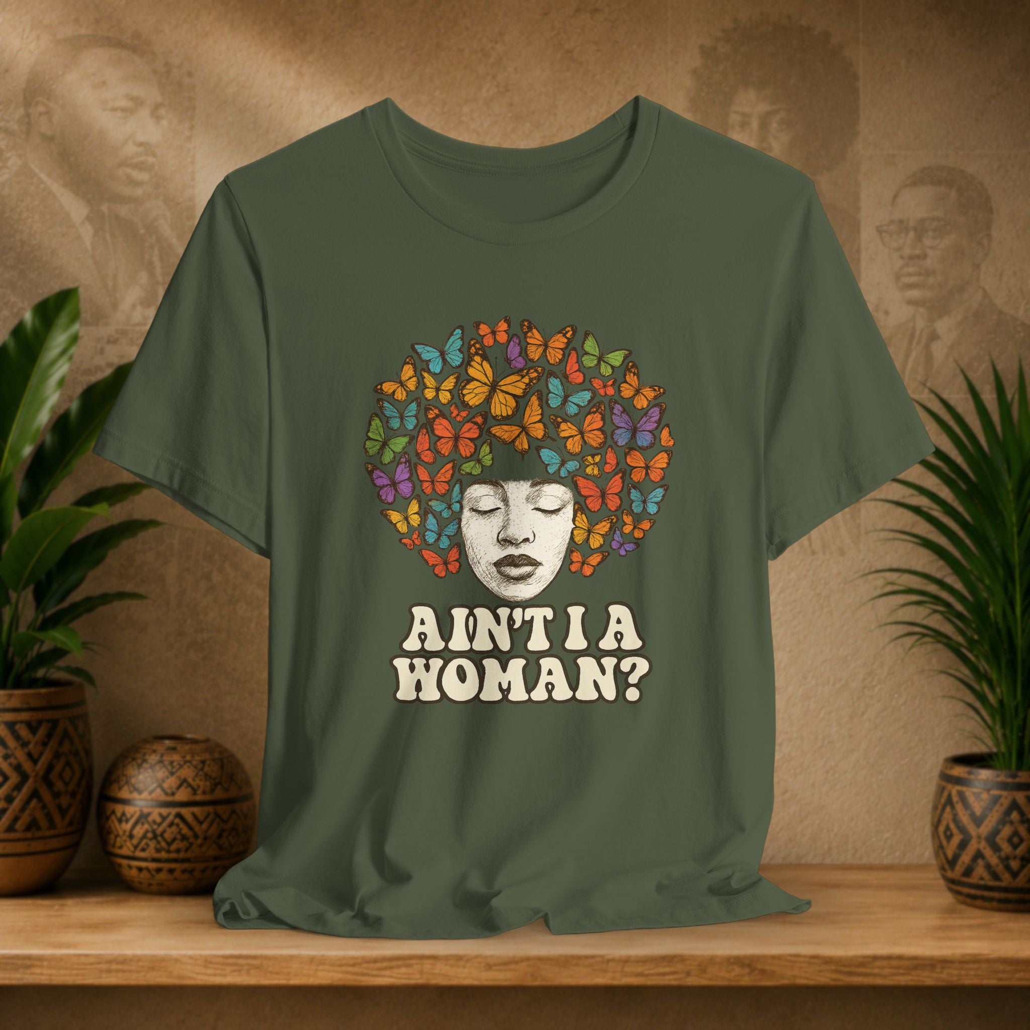 Green t-shirt with a colorful butterfly design and text on a wooden surface with plants and a textured wall in the background.