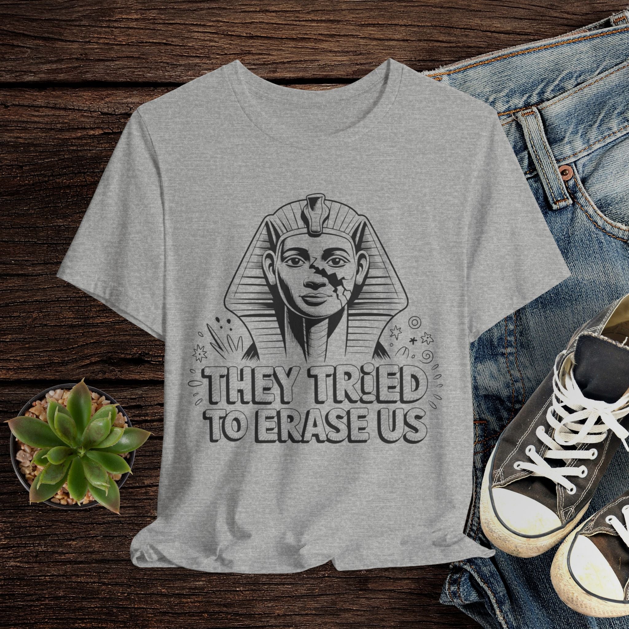Gray t-shirt with Sphinx graphic and text 'They tried to erase us' on a wooden surface with jeans and sneakers.