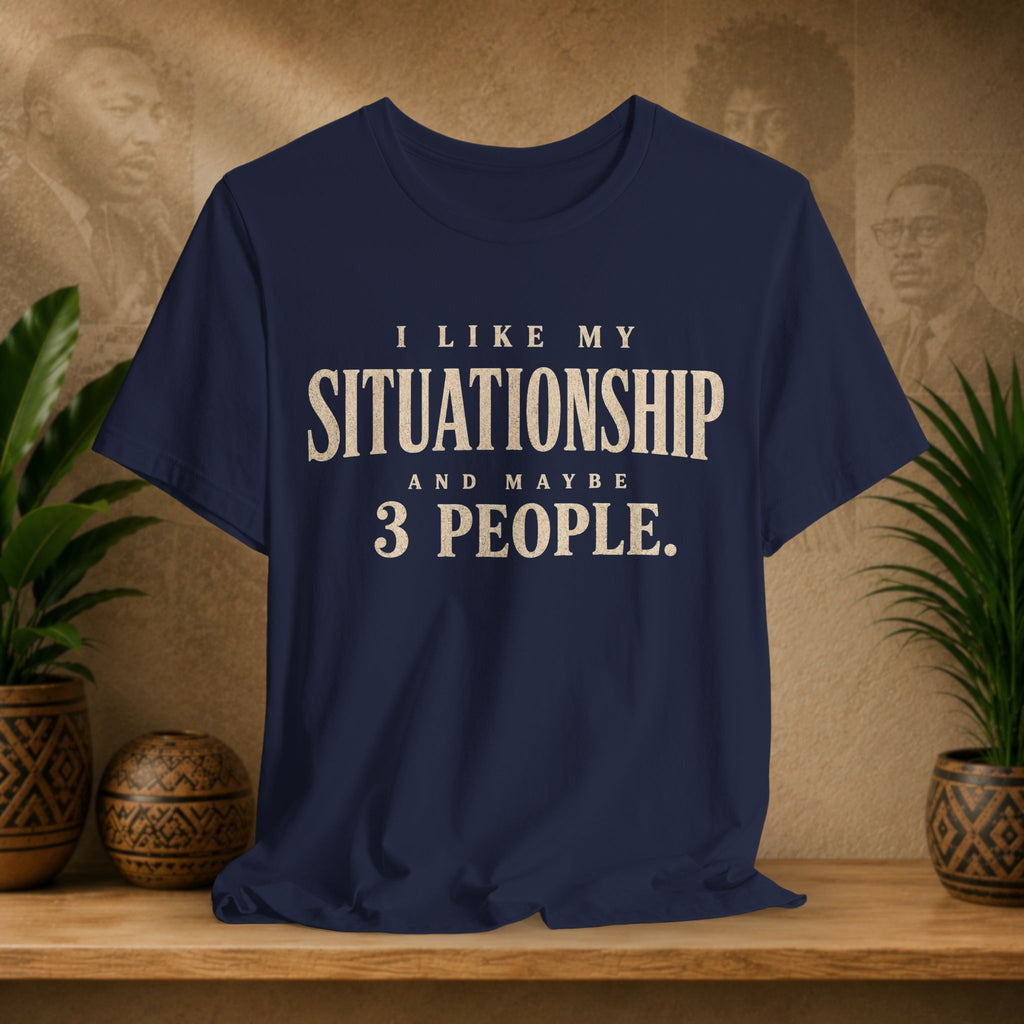 A navy blue graphic t-shirt with cream-colored text reading 'I LIKE MY SITUATIONSHIP AND MAYBE 3 PEOPLE.' displayed on a wooden shelf with potted plants and a textured background.