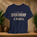 A navy blue graphic t-shirt with cream-colored text reading 'I LIKE MY SITUATIONSHIP AND MAYBE 3 PEOPLE.' displayed on a wooden shelf with potted plants and a textured background.