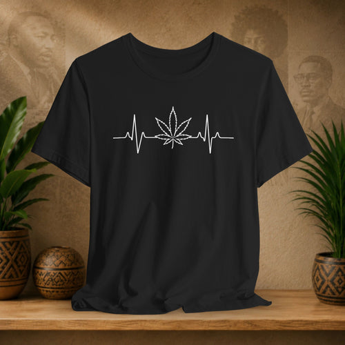 Black t-shirt with a white cannabis leaf and heartbeat line design on a wooden surface with plants and a textured wall background.