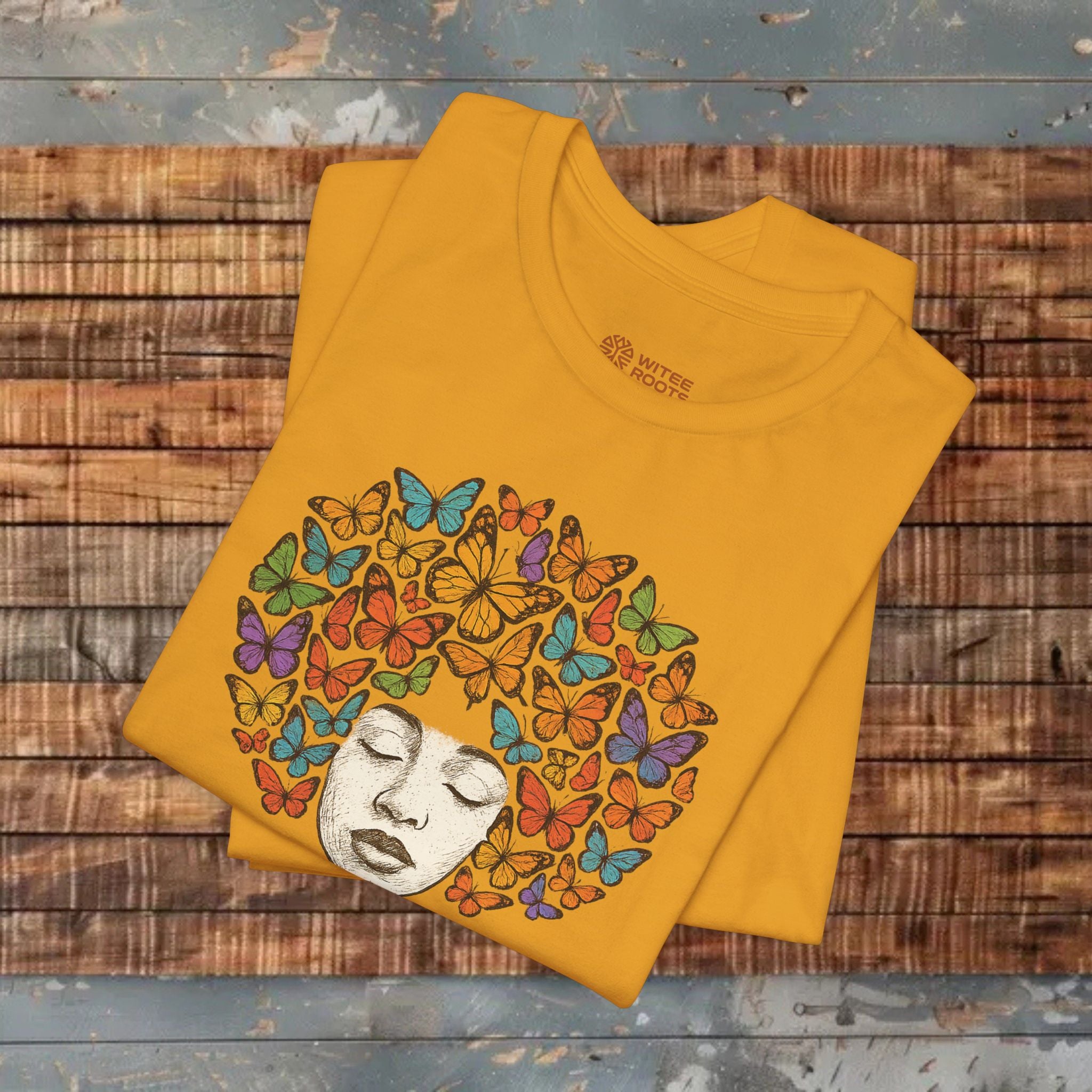 A folded mustard yellow t-shirt with a graphic of a woman's face surrounded by colorful butterflies, displayed on a rustic wooden plank background.