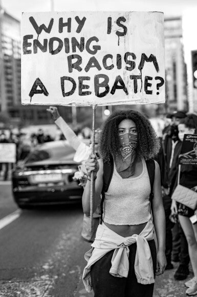 Person holding a protest sign asking 'Why is ending racism a debate?' in an urban setting.