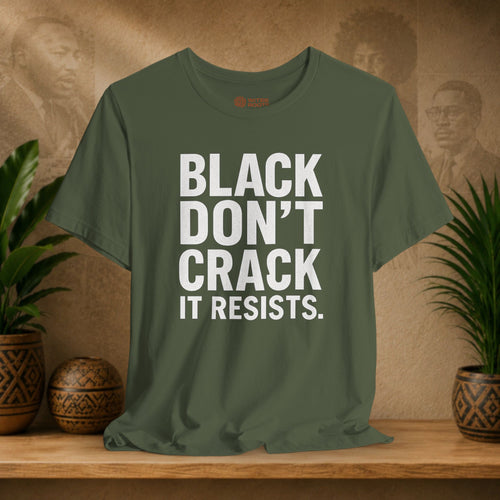 Green t-shirt with 'BLACK DON'T CRACK IT RESISTS.' text on a wooden surface with plants and a textured wall background.