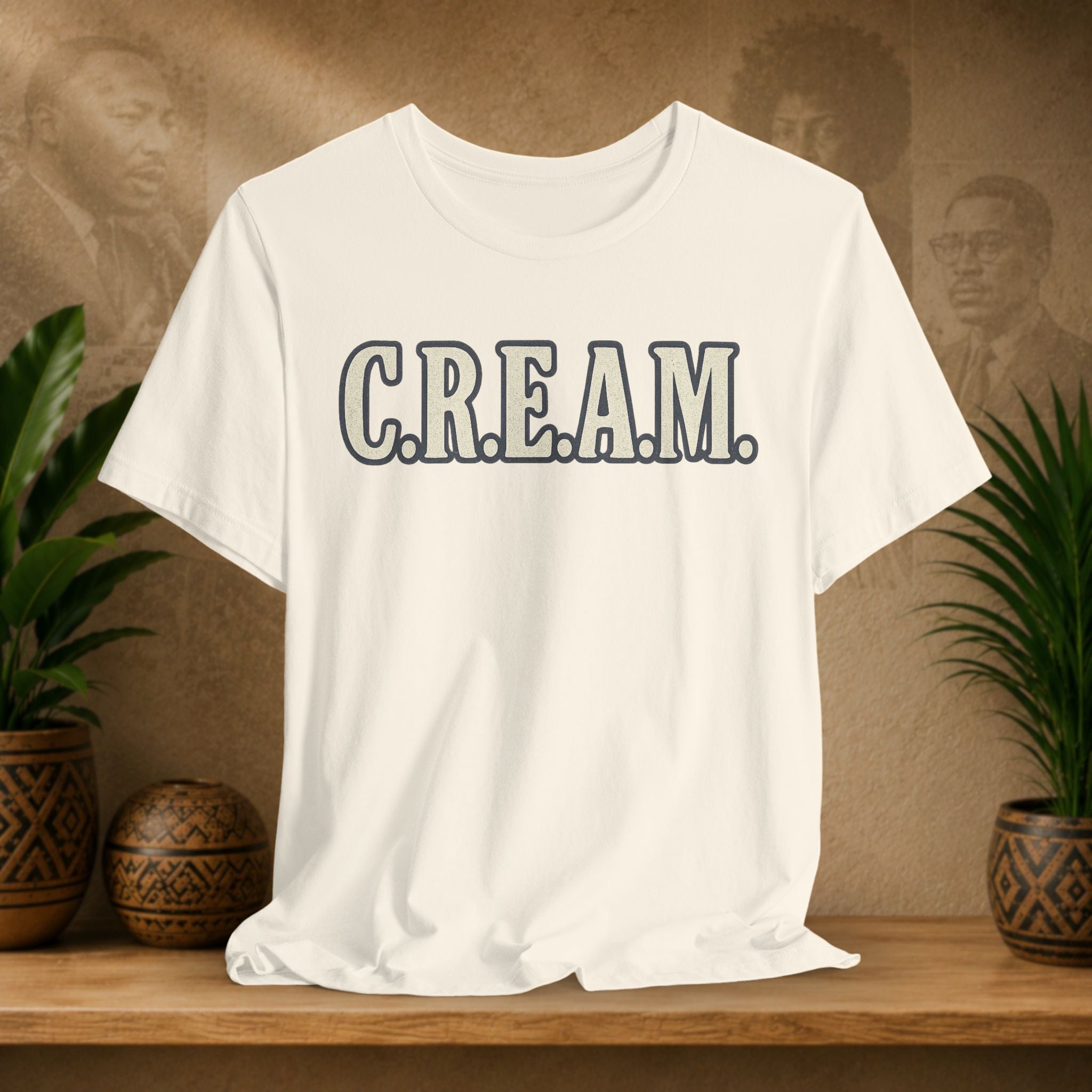 Beige t-shirt with 'C.R.E.A.M.' text on a wooden surface with plants and a textured wall background