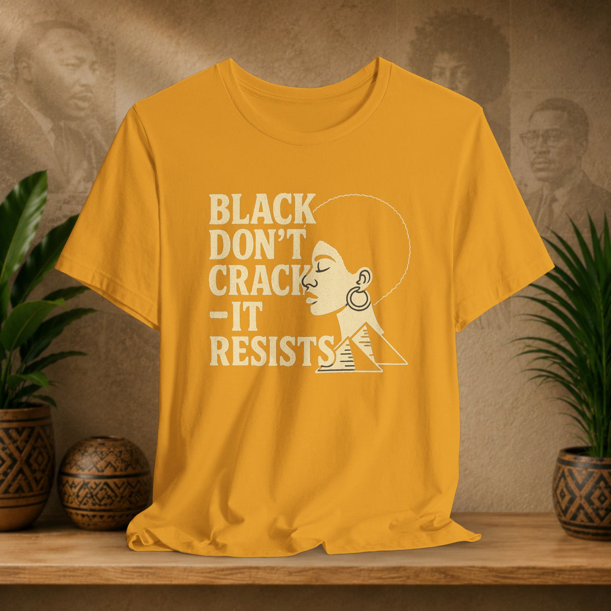 A mustard yellow graphic t-shirt displayed on a wooden shelf. The shirt features a white screen-printed design with the text 'BLACK DON'T CRACK - IT RESISTS' alongside a stylized profile of a woman with an afro and geometric pattern.