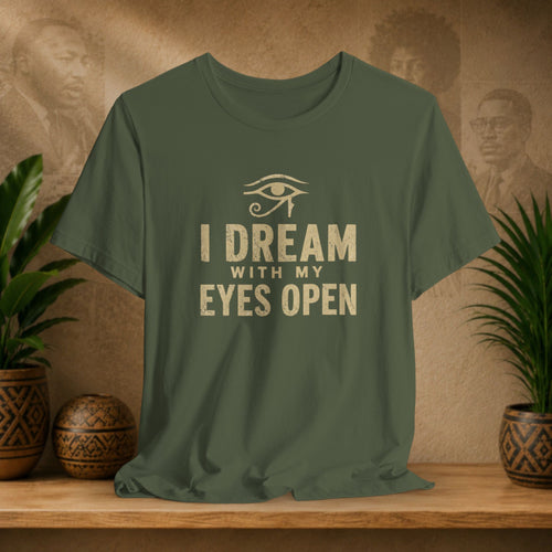 Green t-shirt with 'I Dream with My Eyes Open' text and eye symbol on a wooden surface with plants and wall art.