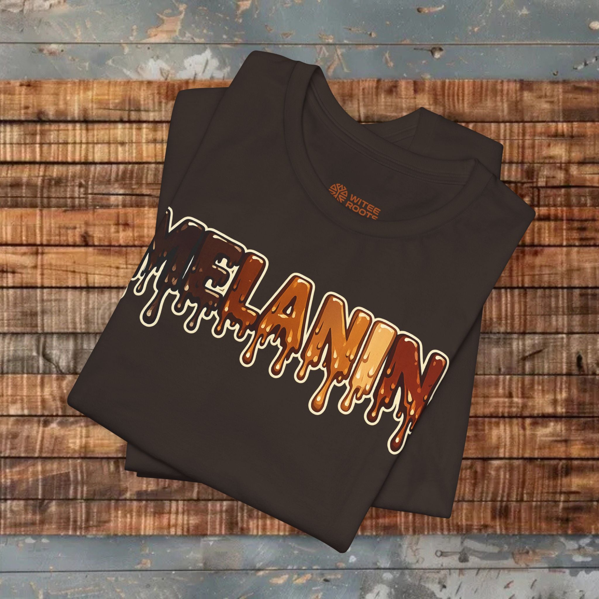 A folded brown t-shirt with a dripping 'MELANIN' graphic in orange, brown, and white, displayed on a rustic wooden plank surface.