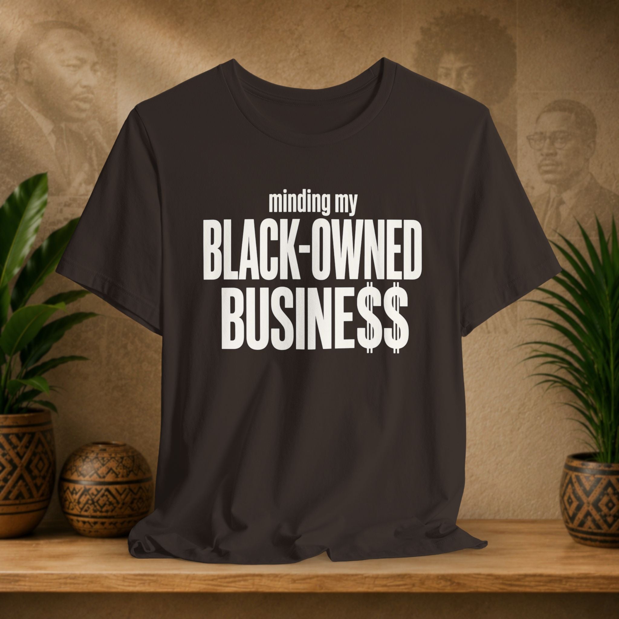 A brown t-shirt with bold white text reading 'minding my BLACK-OWNED BUSINESS$$' displayed on a wooden shelf with potted plants and a textured background.