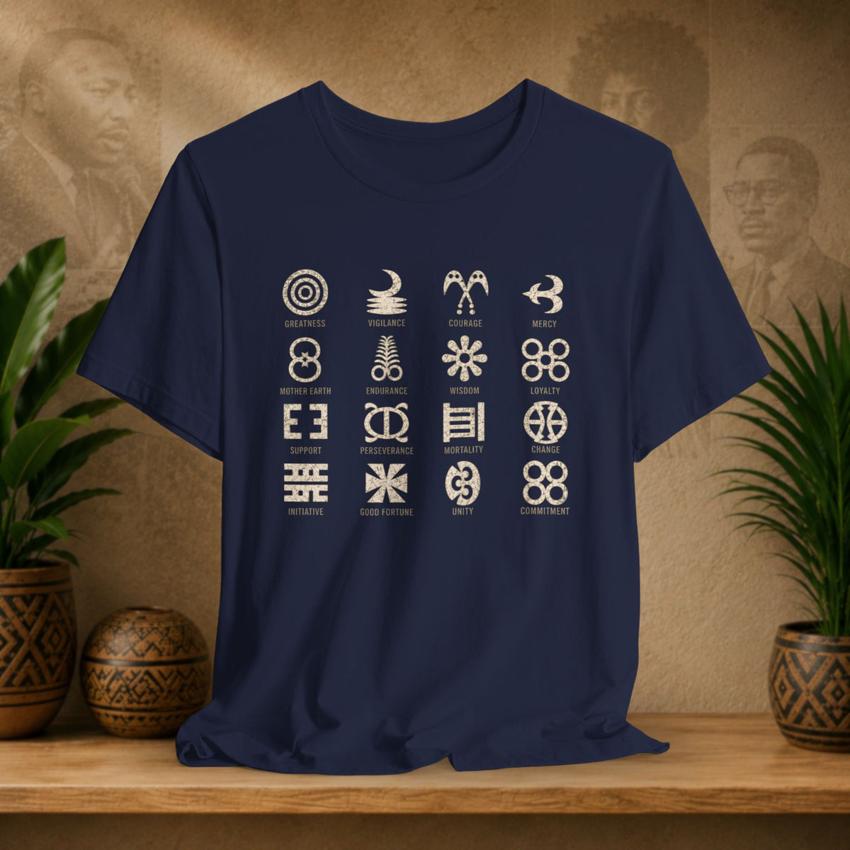 A navy blue short-sleeve t-shirt with a 4x4 grid of white tribal symbols and names printed on the front, displayed on a wooden shelf with potted plants and a textured background.