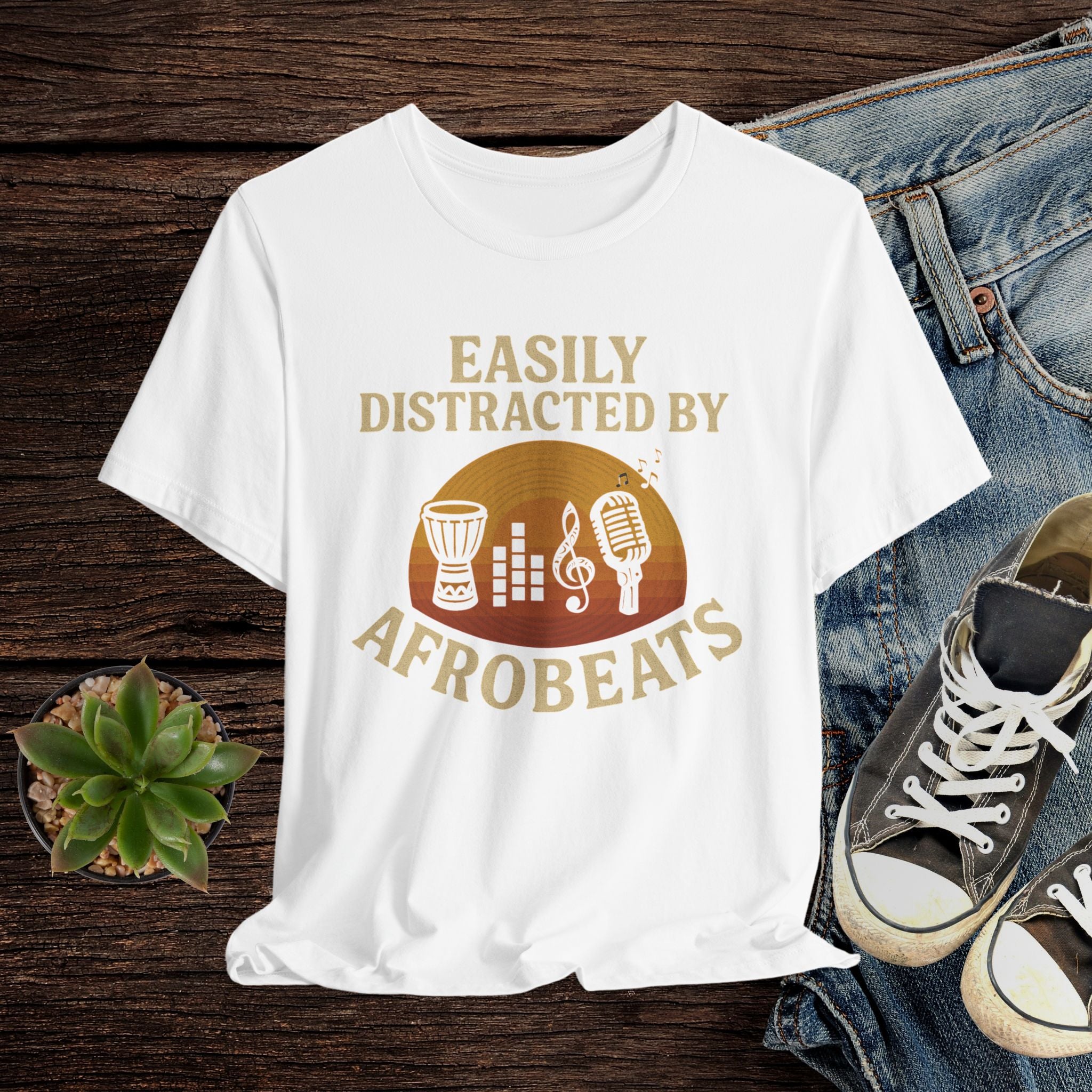 White t-shirt with 'Easily Distracted by AfroBeats' design on a wooden surface with jeans and sneakers.