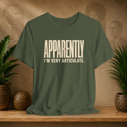 Green t-shirt with 'APPAEARTLY I'M VERY ARTICULATE' text on a wooden surface with plants and a textured wall background.