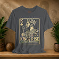 Gray t-shirt with a graphic of a king holding a sword and the text 'Kings Rise' on a wooden surface with plants.