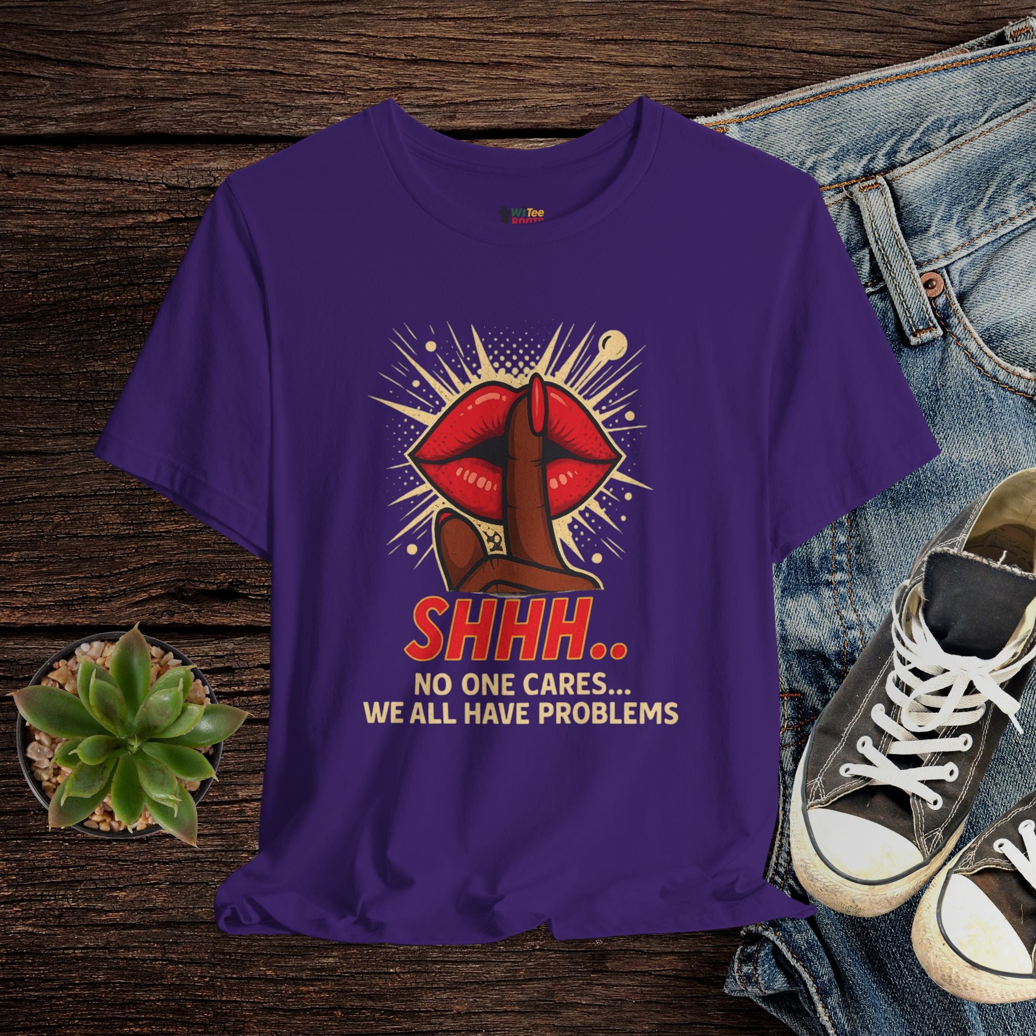 Purple t-shirt with graphic design and text on a wooden surface with jeans and sneakers.