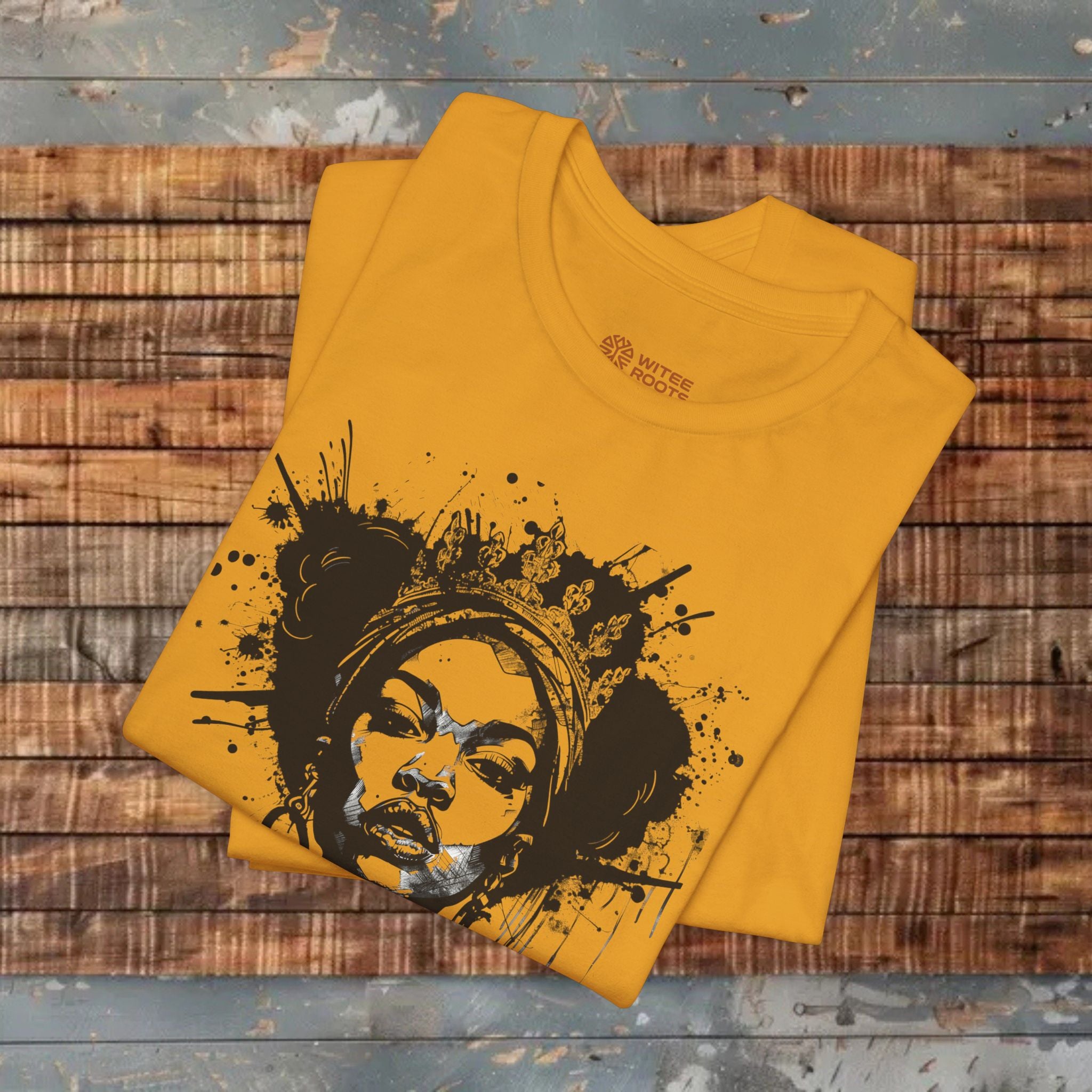 A folded mustard yellow t-shirt with a black and silver graphic of a woman's face surrounded by paint splatters, displayed on a rustic wooden plank background.