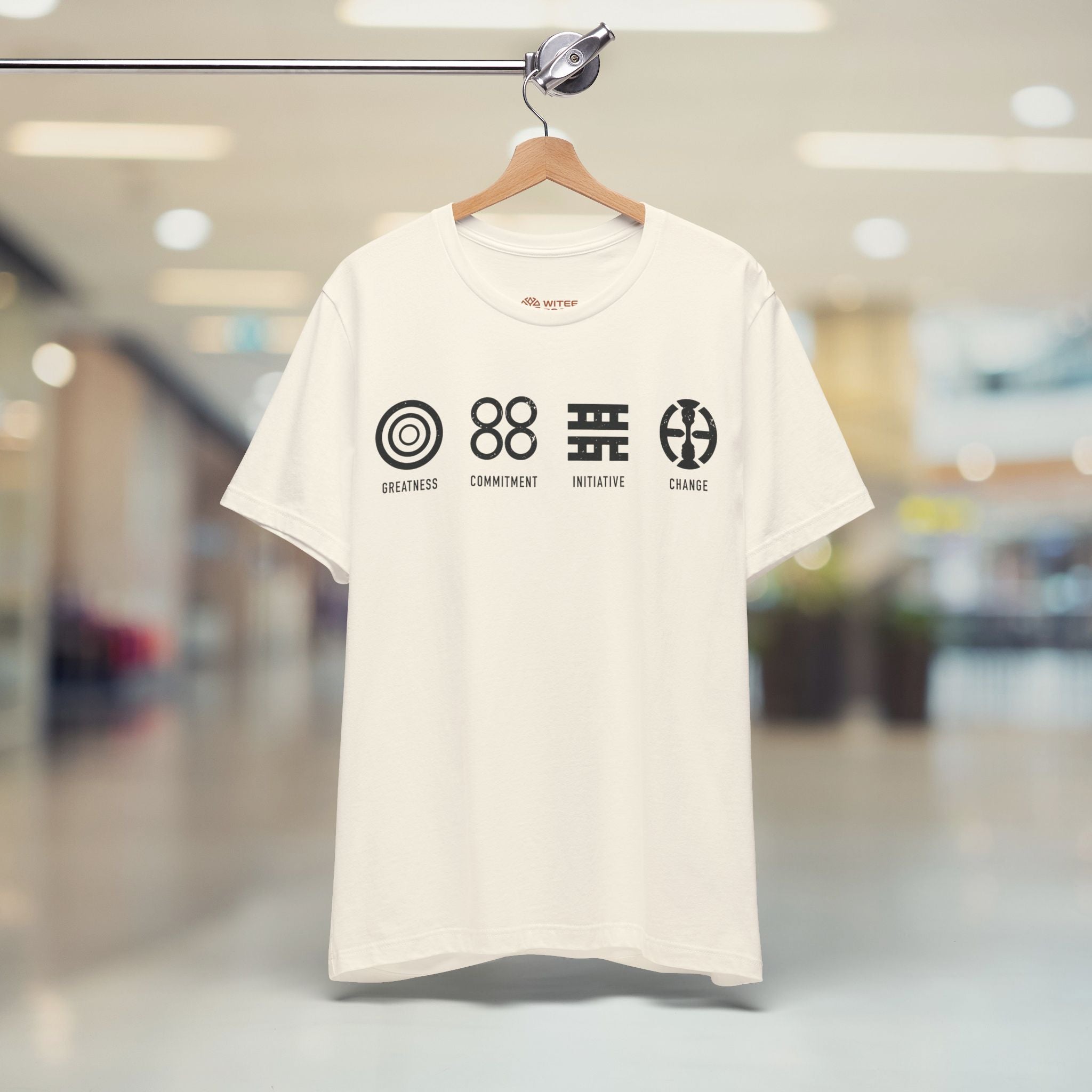 Beige t-shirt with Adinkra symbols on a hanger in a blurred indoor setting 