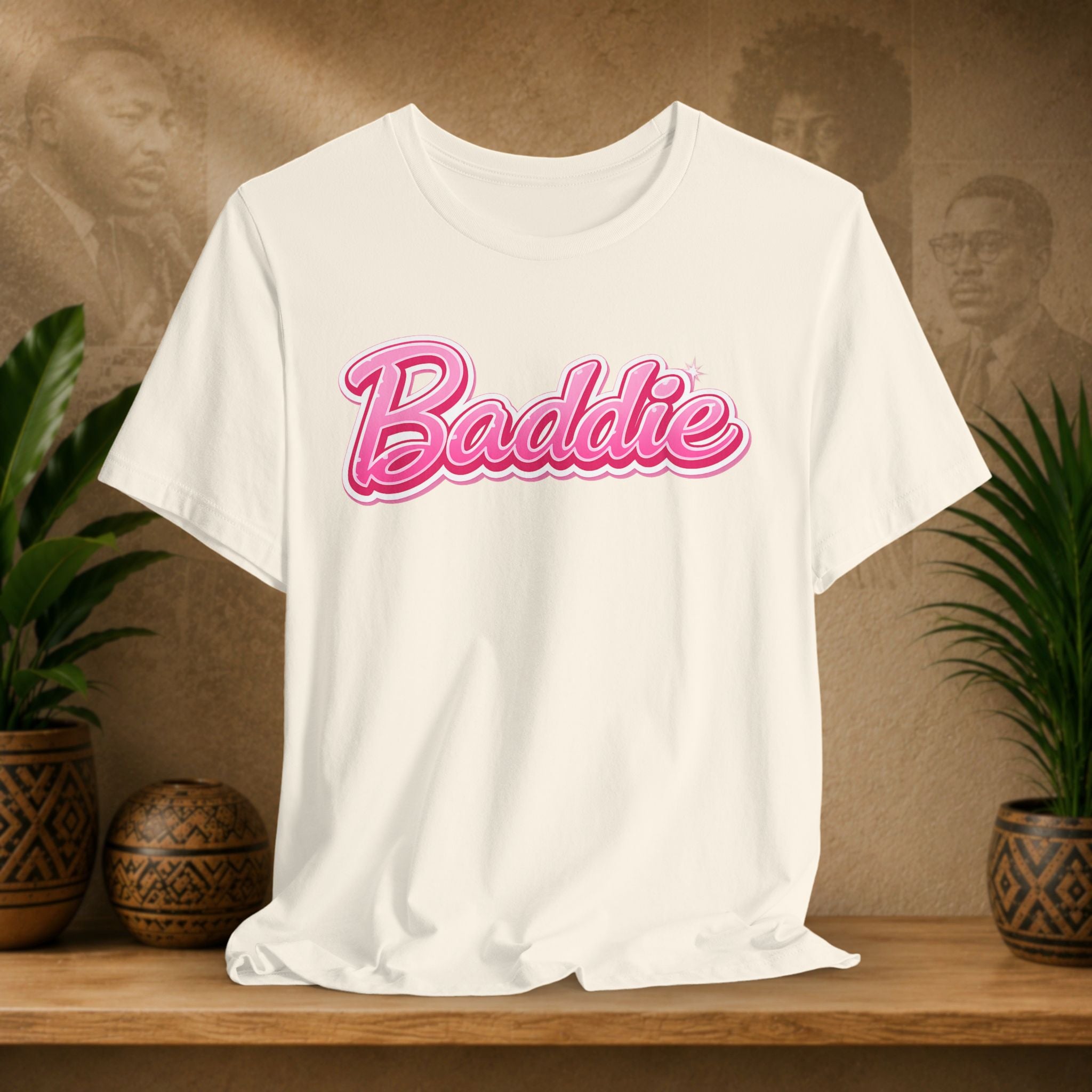 A beige short-sleeve T-shirt with the word 'Baddie' printed across the chest in a pink cursive font with a darker pink outline, displayed on a wooden surface with potted plants and a textured background.